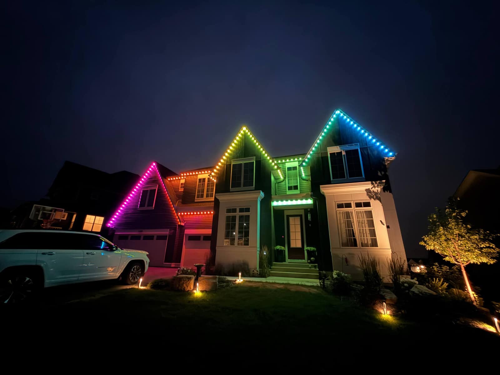 Two-story home at night with multicolor lights — magenta, warm white, green, and blue — following each gable peak and the front porch
