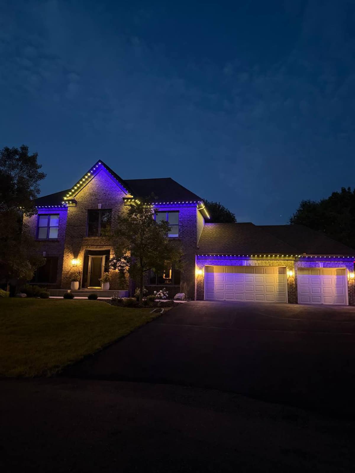 Brick two-story home at dusk with blue and warm white lights following the rooflines and the three-car garage