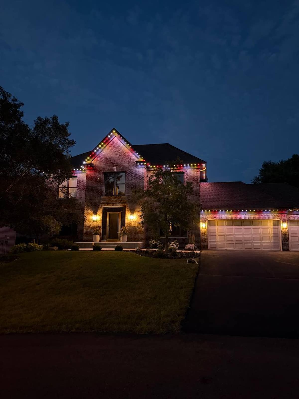 Brick two-story home at dusk with multicolor lights along every roofline and warm porch sconces