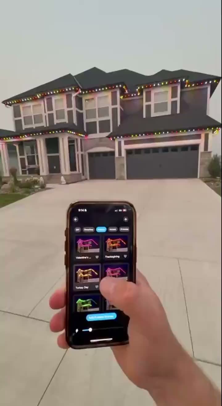 A hand holding a phone running the lighting app, with the home's roofline behind it changing color as the scenes are tapped